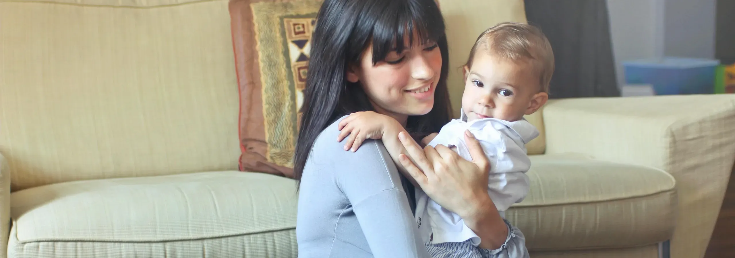 A young au pair lifting a baby in her arms in front of a cream-coloured sofa