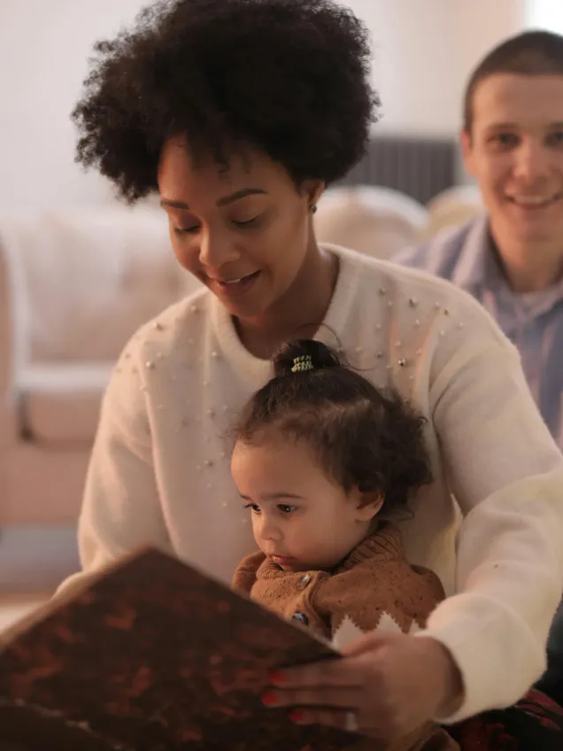 A woman reading a picture book to a toddler on her lap while a man smiles beside them