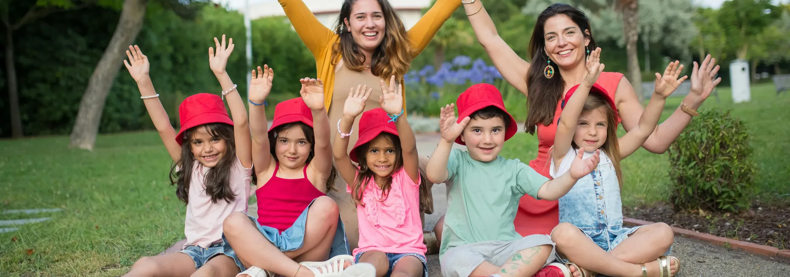 Two women and a group of children in red hats cheering with raised arms in a sunny park