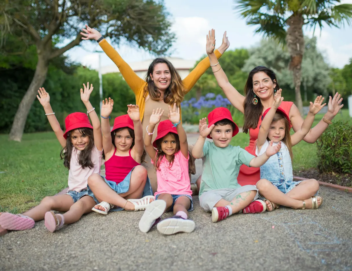 Two women and a group of children in red hats cheering with raised arms in a sunny park